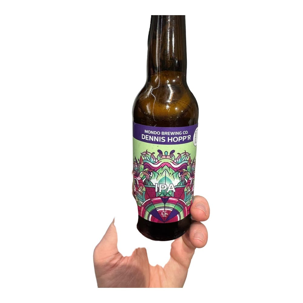A hand holding a brown glass bottle of Dennis Hopp'r IPA beer by Mondo Brewing Co., with colourful label artwork, in an indoor setting with people in the background.