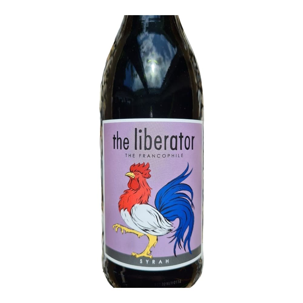 A dark wine bottle with a purple label featuring a colourful rooster illustration, displayed on a bed of white and grey pebbles in natural sunlight.