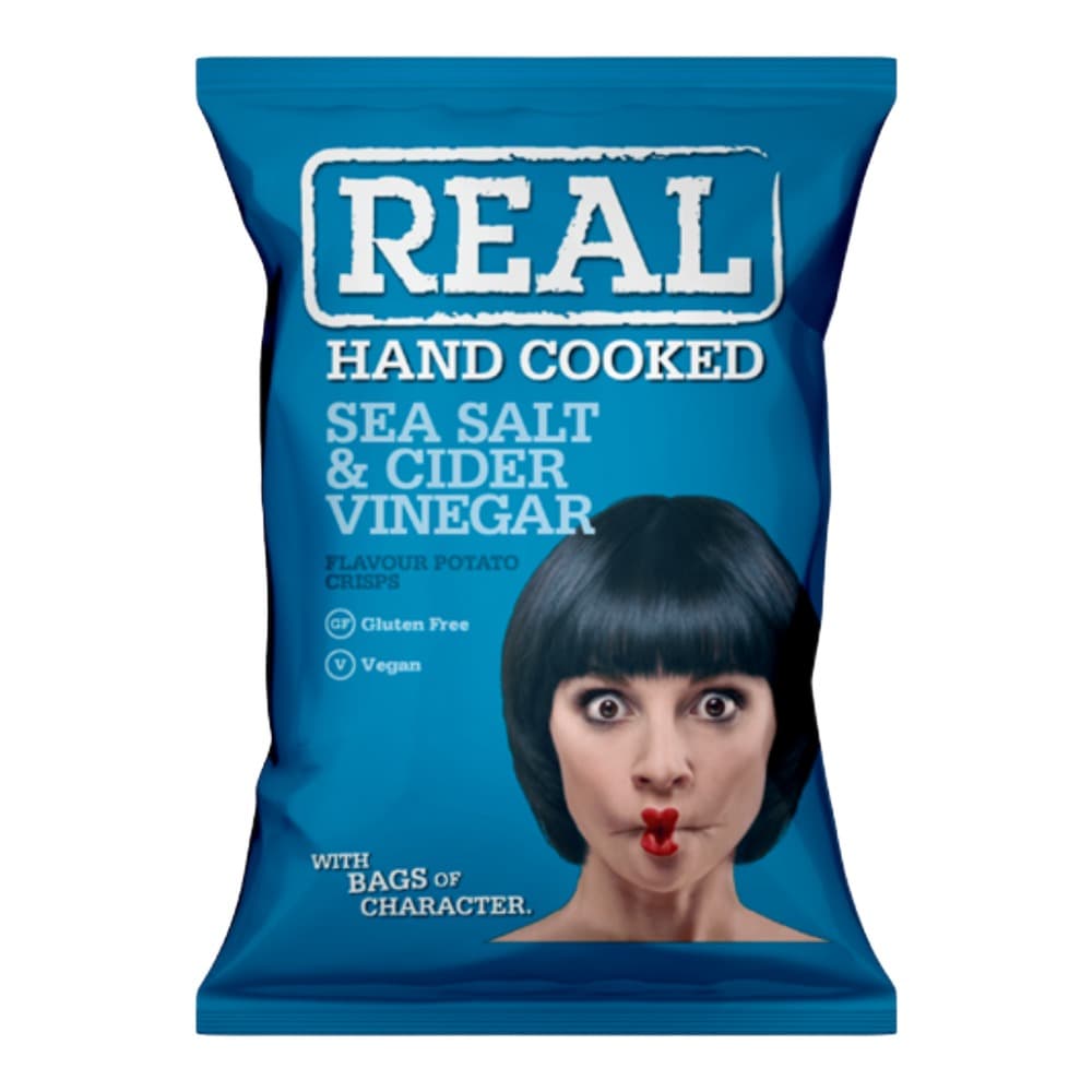 Blue packet of REAL brand hand cooked potato crisps with sea salt and cider vinegar flavour, featuring a woman's face with black bob hair and red lipstick, gluten free and vegan badges visible.