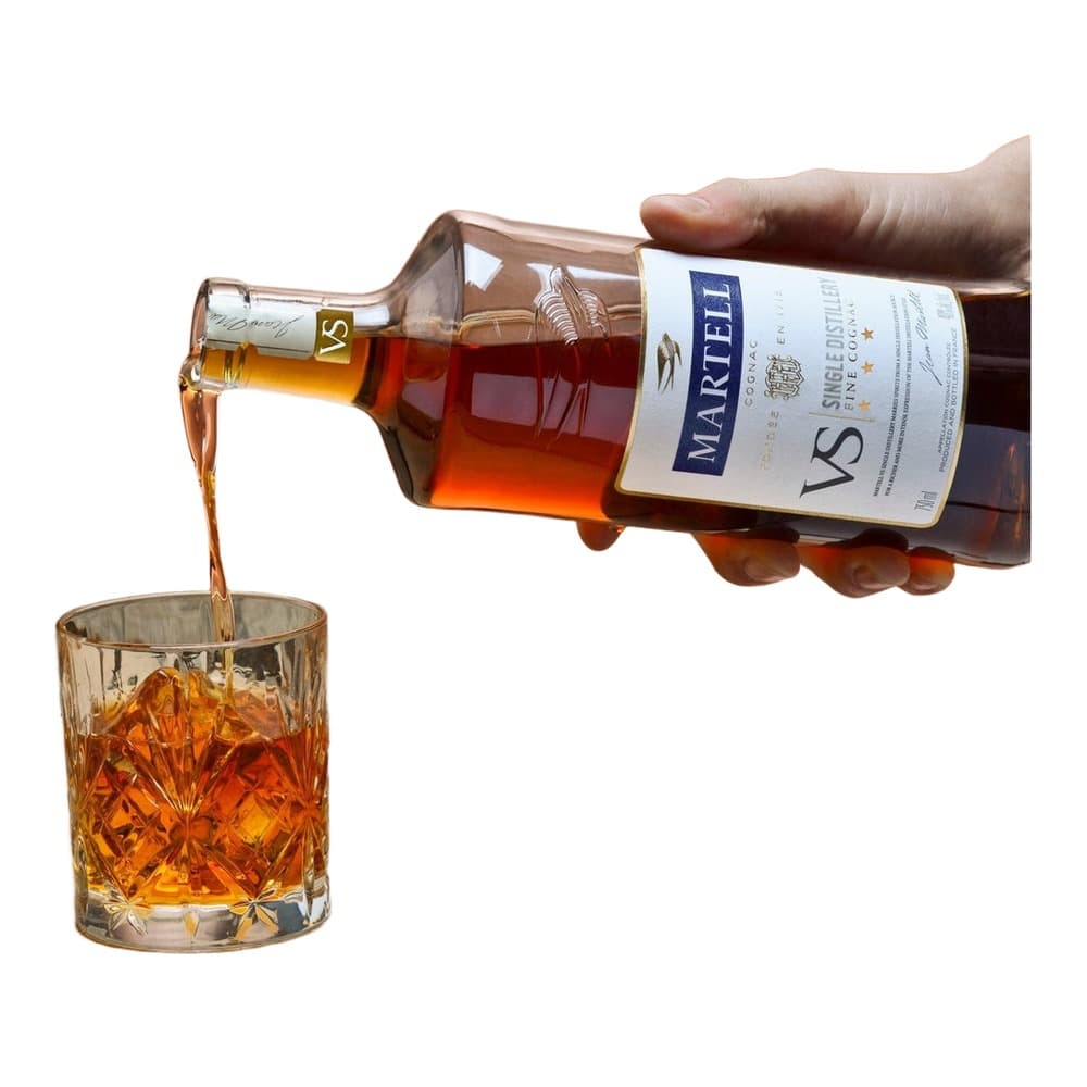 A hand pouring Martell VS Cognac from a bottle into a crystal glass on a wooden barrel with coral background