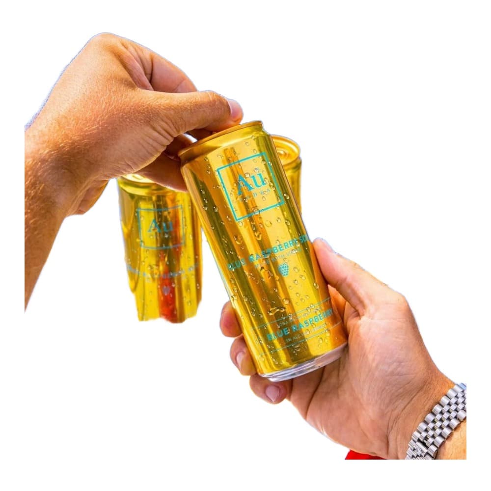 A person's hands holding a golden yellow beverage can with condensation, against a bright blue background with yellow railings and fresh red raspberries visible below.