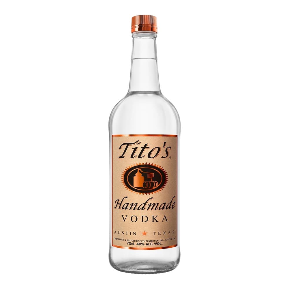 A clear glass bottle of Tito's Handmade Vodka from Austin, Texas, featuring a tan and copper label with a distillery logo, displayed against a white background.