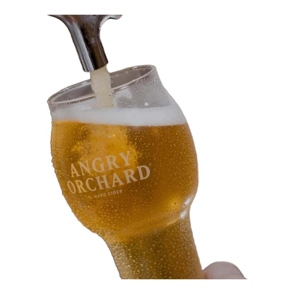 A glass of Angry Orchard hard cider being poured from a bottle, showing the golden liquid and white foam head, with a blurred bar setting in the background