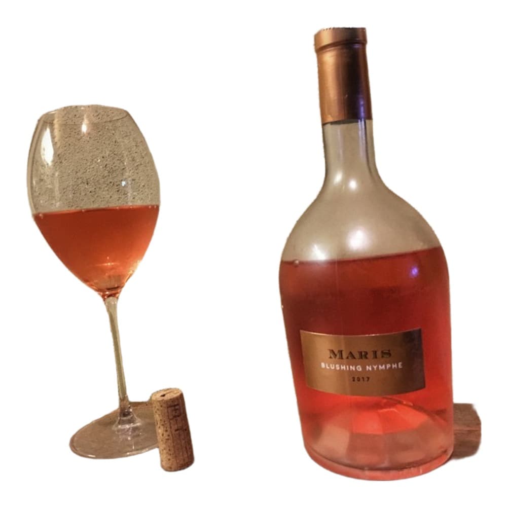 A bottle of Maber Blushing Nymphe rosé wine displayed with a filled wine glass and cork on a textured beige surface.