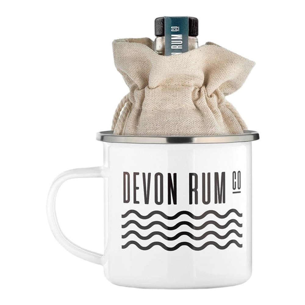 White enamel camping mug branded with Devon Rum Co logo and wave design, topped with a cream linen pouch containing a dark glass rum bottle with navy label.
