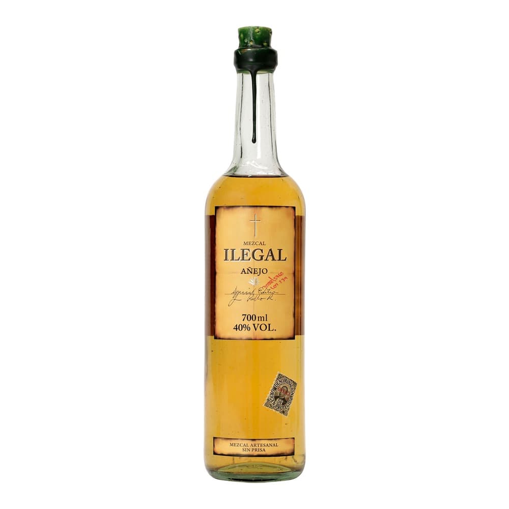 A 700ml bottle of Ilegal Mezcal Añejo with a golden-amber liquid, green capsule top, and cream-coloured label featuring a cross and handwritten signature against a white background.