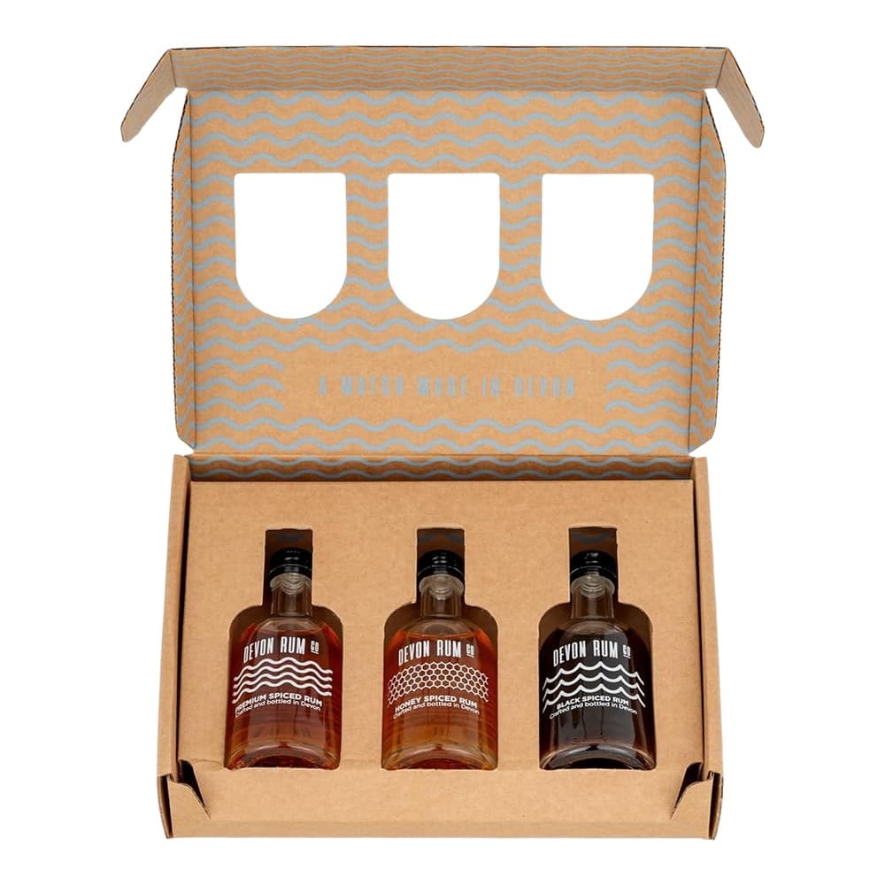 Open cardboard gift box containing three Devon Rum Co spiced rum bottles with black caps, displayed on kraft paper padding with a decorative wavy pattern insert lid.