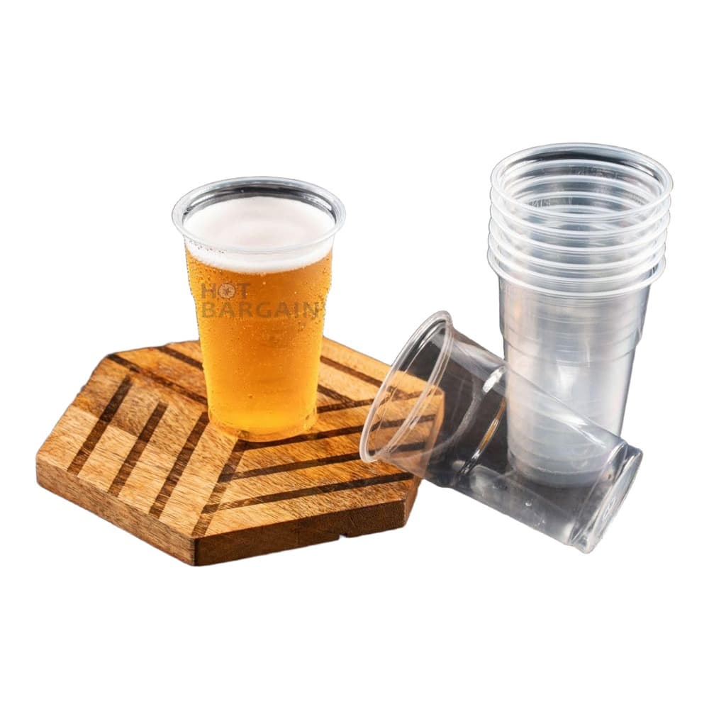A pint glass of golden beer with a white foam head sits on a wooden serving board alongside empty clear plastic cups, set against a dark textured background.