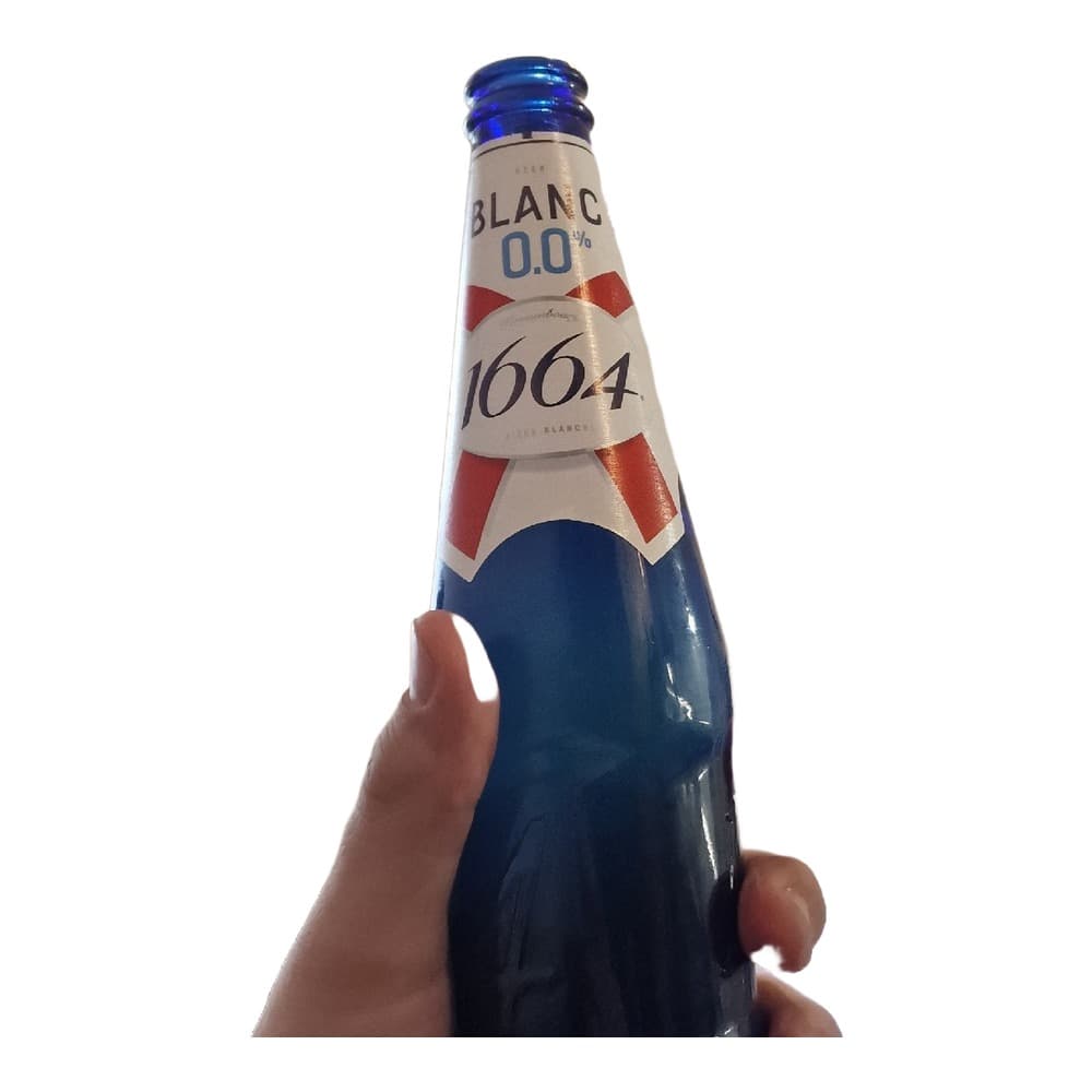 A hand holding a blue glass bottle of Kronenbourg 1664 Blanc alcohol-free beer with white and red label against an indoor background.